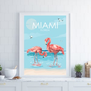 K&ouml;nnte beinhalten: Ein gerahmtes Poster mit blauem und wei&szlig;em Hintergrund, auf dem zwei rosa Flamingos im Wasser stehen. Das Poster zeigt oben "Miami, Florida" an.
