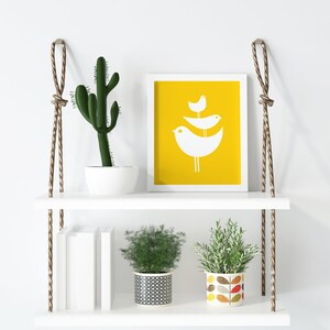 May include: White floating shelves with rope hangers, featuring a cactus in a white pot, a framed print with a yellow background and white birds, a stack of books, and two potted plants. One plant is in a black and white patterned pot, and the other is in a colorful pot with a leaf design.
