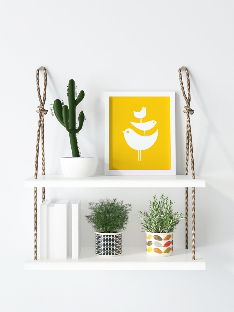 May include: White floating shelves with rope hangers, featuring a cactus in a white pot, a framed print with a yellow background and white birds, a stack of books, and two potted plants. One plant is in a black and white patterned pot, and the other is in a colorful pot with a leaf design.