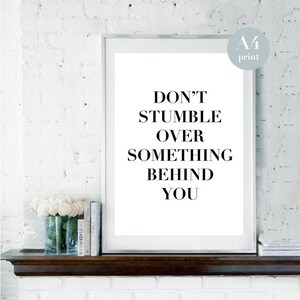 May include: A white framed print with the text "Don't stumble over something behind you" in black lettering. The print is on a white brick wall with a wooden shelf below it.