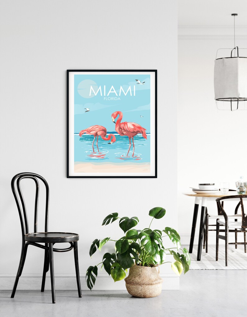 K&ouml;nnte beinhalten: Ein Poster mit blauem Hintergrund und dem Text "Miami, Florida" in Wei&szlig;. Zwei rosafarbene Flamingos stehen im Wasser mit einem wei&szlig;en Sandstrand im Hintergrund.