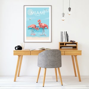 K&ouml;nnte beinhalten: Ein gerahmter Druck mit blauem Himmel und Meereshintergrund, auf dem zwei rosafarbene Flamingos im Wasser stehen. Der Druck tr&auml;gt oben den Text "MIAMI FLORIDA".