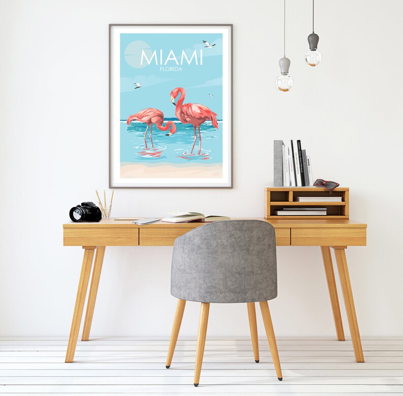 K&ouml;nnte beinhalten: Ein gerahmter Druck mit blauem Himmel und Meereshintergrund, auf dem zwei rosafarbene Flamingos im Wasser stehen. Der Druck tr&auml;gt oben den Text "MIAMI FLORIDA".