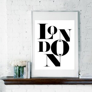 May include: Black and white typography print with the word "LONDON" in a bold, stylized font. The print is framed in a simple silver frame and displayed on a white brick wall.