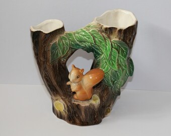 May include: A ceramic vase shaped like a tree trunk with a squirrel. The vase has two openings at the top and a hole in the middle. The squirrel is brown and orange, and the leaves are green.
