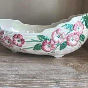 Puede incluir: Un macetero de cerámica blanca con un borde festoneado y un diseño floral. El macetero está decorado con flores rosas y hojas verdes.