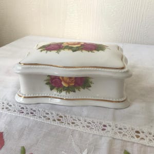 May include: White porcelain trinket box with a gold trim and a floral design featuring pink, yellow, and green roses.