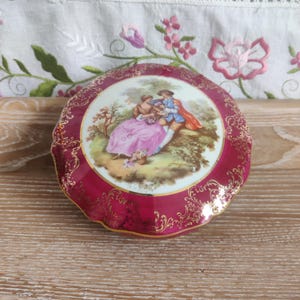 May include: A round, red trinket box with gold trim and a painted scene of a couple in a garden. The box has a scalloped edge and a white interior.