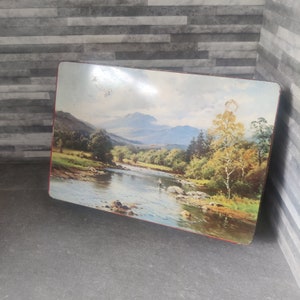 May include: A vintage tin box with a scenic image of a river flowing through a valley with mountains in the background. The river is surrounded by trees and rocks. A single figure is fishing in the river.
