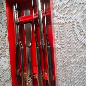 Puede incluir: Un juego de seis cuchillos plateados en una caja roja. Los cuchillos tienen mangos decorativos y varios estilos de hoja, incluido un cuchillo de mantequilla. La caja está abierta, revelando los cuchillos dispuestos en fila.