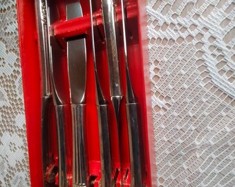 Puede incluir: Un juego de seis cuchillos plateados en una caja roja. Los cuchillos tienen mangos decorativos y varios estilos de hoja, incluido un cuchillo de mantequilla. La caja está abierta, revelando los cuchillos dispuestos en fila.