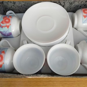 May include: A white porcelain tea set with six cups, saucers, and a serving tray. Each cup features a floral and butterfly design in red, orange, and blue. The set is neatly arranged inside a decorative box.