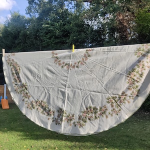 May include: A large, cream-coloured tablecloth with a floral pattern of pink and green flowers. The tablecloth is hanging on a washing line in a garden. The tablecloth is round and has a scalloped edge.