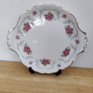 Puede incluir: Un plato de servir de porcelana blanca con un borde festoneado y un borde dorado. El plato está decorado con un patrón floral de rosas rosas y hojas verdes sobre un fondo gris.