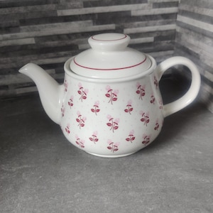May include: White ceramic teapot with a lid, decorated with a pattern of small pink flowers and green leaves. The teapot has a curved spout and a rounded handle. A thin red line accents the lid and the rim.