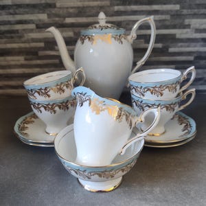 Op de afbeelding: Een theeservies van wit porselein met een lichtblauwe band en sierlijke gouden bladmotieven. De set bevat een theepot met deksel, vier theekopjes met bijpassende schoteltjes, een melkkannetje en een suikerpot, allemaal met gouden randen.