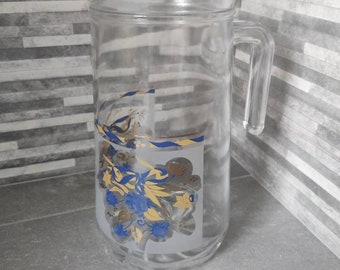 May include: A clear glass pitcher with a floral design in blue, yellow, and gold. The pitcher has a lid and a handle.