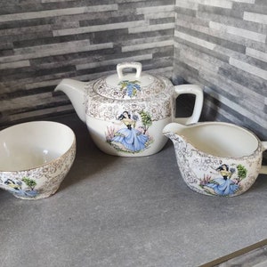 May include: A vintage tea set with a white background and gold accents. The teapot, creamer, and sugar bowl feature a floral design with a woman in a blue dress.