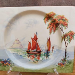 May include: Rectangular decorative plate with a painted seascape scene. The plate features a body of water with sailboats, a small island, and a tree with orange blossoms. The plate has a cream-coloured background and a green border.