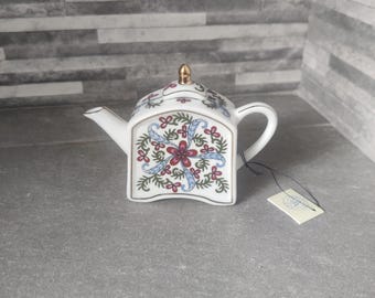Puede incluir: Una pequeña tetera de cerámica blanca con un adorno y asa dorados. La tetera presenta un diseño floral en rojo, azul y verde. Una pequeña etiqueta está sujeta al asa. La tetera mide aproximadamente 13 cm de largo.