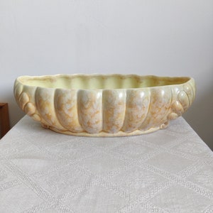 May include: A large, oval-shaped ceramic planter with a light yellow glaze and a brown marbled pattern. The planter has a scalloped edge and decorative details on the sides.