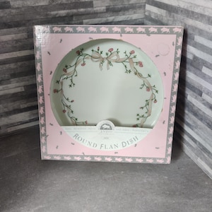 Könnte beinhalten: Eine weiße runde Flan-Form mit einem rosafarbenen Blumendesign und einem grünen Band. Die Form befindet sich in einer rosa und weißen Schachtel mit der Aufschrift "Round Flan Dish" auf der Vorderseite.