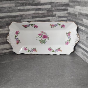 Puede incluir: Una bandeja rectangular blanca para servir con un borde dorado y un diseño floral. La bandeja tiene un borde festoneado y presenta flores rosas y moradas con hojas verdes.