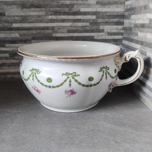 May include: A white porcelain chamber pot with a gold rim and a decorative handle. The pot is decorated with green garland and pink rose accents. A vintage item.