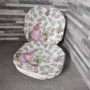 May include: A stack of vintage square plates with scalloped edges. Each white plate features a gold floral design and a central illustration of a woman in a pink dress. The plates have gold trim.