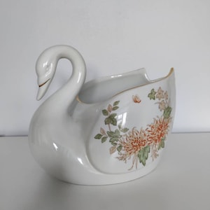 May include: White porcelain swan planter with a floral design featuring orange and green chrysanthemums and leaves. The swan's neck is curved and the planter has a wide, open mouth.