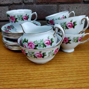 May include: A classic tea set comprising white porcelain cups, saucers, a creamer, and a sugar bowl. Each item is decorated with pink rose floral designs and gold edging. The set includes several cups and saucers, ideal for serving tea.