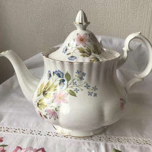 Puede incluir: Tetera blanca con un diseño floral con flores rosas, azules y verdes. La tetera tiene un borde dorado alrededor de la tapa y la base.
