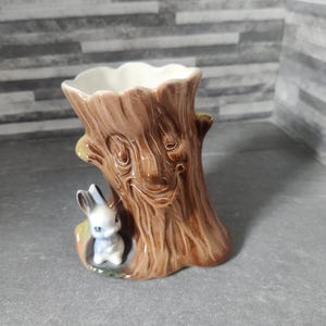 May include: A ceramic vase shaped like a tree trunk, with a brown, textured exterior and a white interior. A small, white and blue rabbit figurine is nestled at the base. The vase is designed for decorative purposes.
