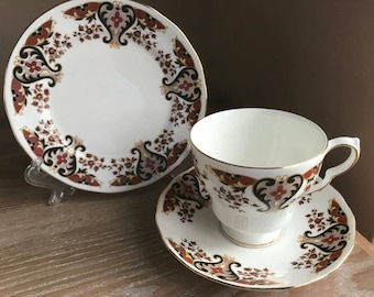 May include: A white teacup and saucer set with a floral pattern in shades of red, brown, and black. The pattern features paisley-like designs and gold accents.