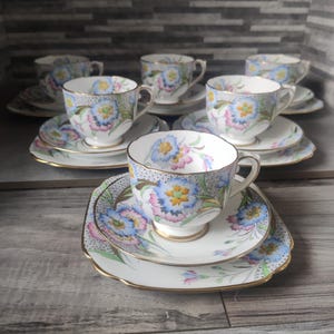 May include: A set of fine china tea cups and saucers featuring a delicate floral pattern with blue, pink, and yellow blossoms on a white background. Each piece is accented with gold trim and a subtle gray stippled texture along the edges.
