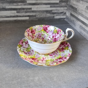 Könnte beinhalten: Ein Blumen-Teetassen- und Untertassen-Set mit einem rosa-violetten Blumenmuster. Tasse und Untertasse sind weiß mit goldener Verzierung.