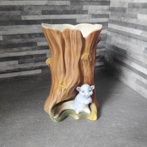Könnte beinhalten: Eine Keramikvase in Form eines Baumstamms, mit hellbrauner Außenseite und weißer Innenseite. Am Fuß befindet sich eine hellblaue Eichhörnchenfigur. Die Vase hat ein einzigartiges, rustikales Design.