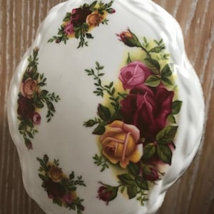 Op de afbeelding: Wit porseleinen sieradendoosje met een bloemenontwerp met rode, gele en roze rozen. Het doosje heeft een gegolfde rand.