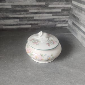 May include: A white ceramic jar with a floral design and a lid shaped like a rose. The jar has a pink, green, and white floral pattern.
