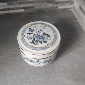 May include: A round, white ceramic trinket box with a lid. The lid and sides feature a floral design in blue and pink, with gold trim. The box is sitting on a gray surface.