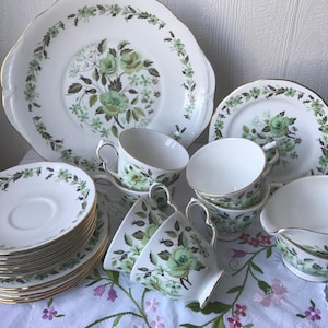 Op de afbeelding: Een vintage set theekopjes en schoteltjes met een bloemenpatroon. De set bevat een theepot, een roomkan, een suikerpot en een grote serveerschaal. De kopjes en schoteltjes zijn wit met een groen-wit bloemenontwerp.
