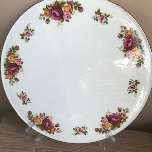 Puede incluir: Un plato de pastel de cerámica blanca con un borde dorado y un diseño floral con rosas rosas, amarillas y verdes. El plato está sobre una superficie de madera.