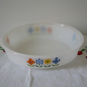 Könnte beinhalten: Weiße Keramik-Backform mit Blumenmuster. Die Form hat einen gewellten Rand und zeigt vier bunte Blumen in den Farben Blau, Gelb, Orange und Rot.