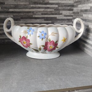 Può includere: Fioriera in ceramica color crema con due manici e bordo smerlato. Decorata con motivi floreali rosa, blu e gialli. La base è rotonda e le maniglie sono arcuate.