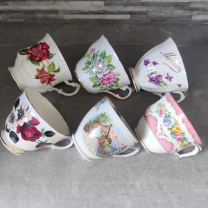 Peut inclure: Six tasses à thé blanches avec des bordures dorées, chacune ornée d'un motif floral différent. Les tasses présentent des fleurs dans des tons de rouge, rose, violet et jaune. Une tasse porte l'inscription "Mother".