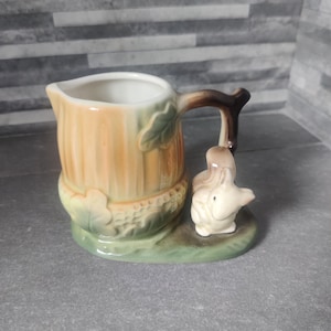 May include: A ceramic creamer with a pumpkin-shaped body in shades of orange and green. A brown handle and a small white squirrel figurine are attached. The creamer sits on a green base.