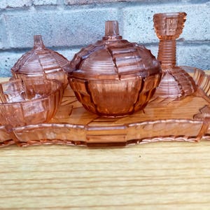 Könnte beinhalten: Ein Vintage-Eitelkeitsset aus rosa Glas, einschließlich einer Ablage, eines abgedeckten Glases, zwei kleiner Schalen und eines Kerzenhalters. Das Set hat ein geometrisches Design und wird auf einer Holzoberfläche präsentiert.