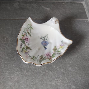 Puede incluir: Un plato de porcelana blanca en forma de concha con un borde dorado. El plato está decorado con un patrón floral de flores rosas, azules y amarillas.