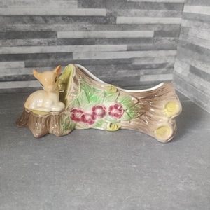 Könnte beinhalten: Ein Keramik-Pflanztopf in Form eines Baumstamms mit einem Reh, das auf einem Baumstumpf sitzt. Der Topf ist mit grünen Blättern, rosa Blüten und brauner Rinde verziert.