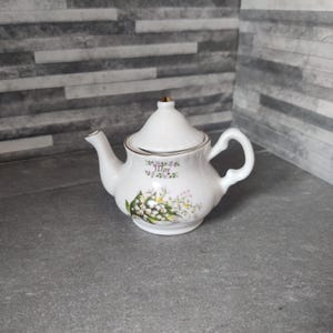 Puede incluir: Una tetera de porcelana blanca con un diseño floral con flores de lirio de los valles. La tetera tiene un borde dorado y dice "Mayo" en la parte delantera.